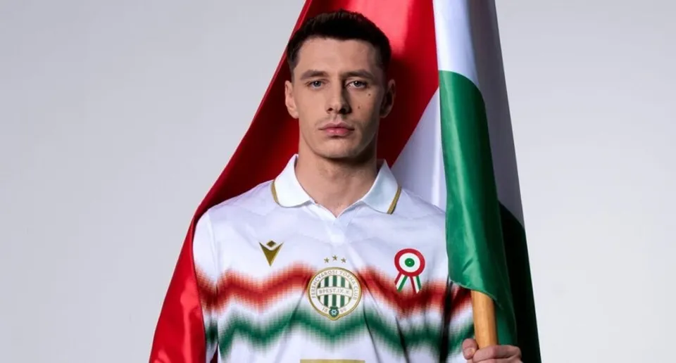 Un bărbat cu părul scurt și cămașă albă cu un logo și un emblema colorată stă în fața unui steag vertical.