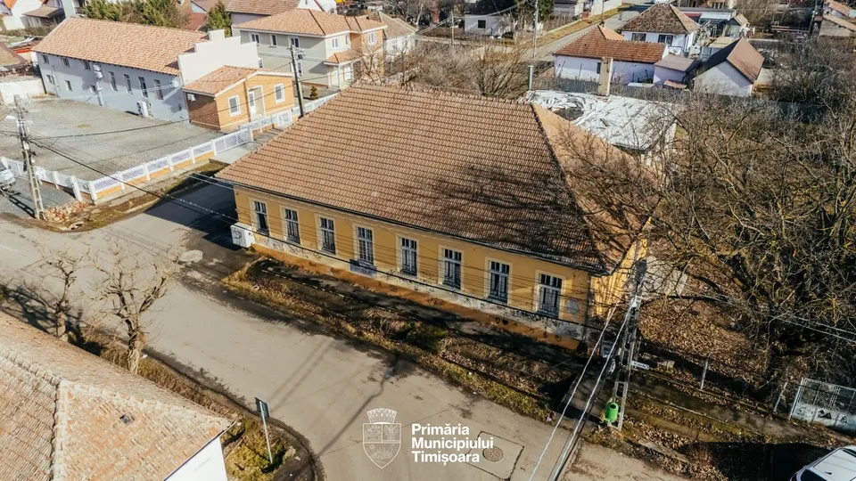 O clădire cu fațadă galbenă și acoperiș de țiglă maro este situată lângă un drum asfaltat, fiind înconjurată de vegetație și alte construcții.
