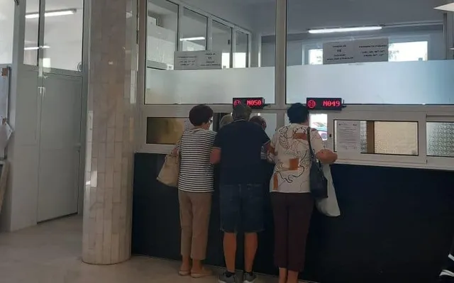 Trei persoane stau în fața unor ghișee într-o încăpere cu pereți albi și stâlpi de marmură.