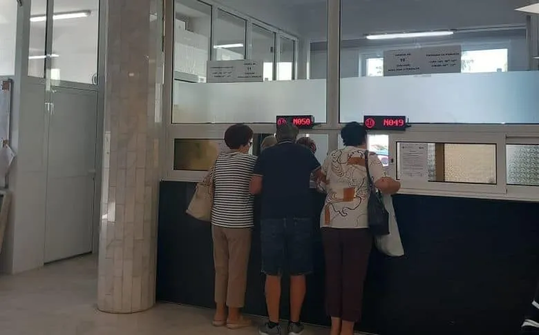Trei persoane stau în fața unor ghișee într-o încăpere cu pereți albi și stâlpi de marmură.