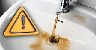 Un robinet de chiuvetă lasă curgere de apă murdară, de culoare maro, într-o chiuvetă albă lângă un semn de avertizare galben.