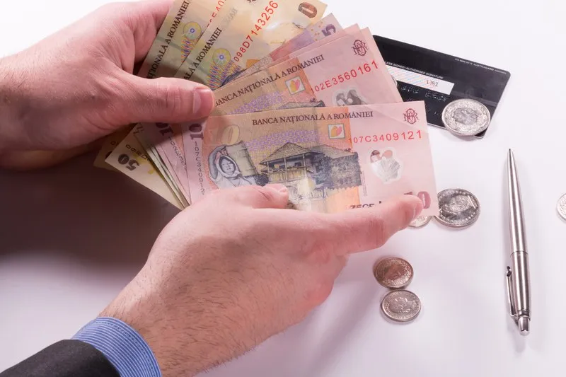 Două mâini țin un teanc de bancnote românești de peste cincizeci de lei pe un fundal alb lângă monede și un pix argintiu.