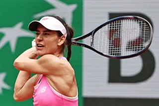 O femeie cu vizieră albă și echipament roz balancează o rachetă de tenis în poziție de lovitură pe un teren de sport.
