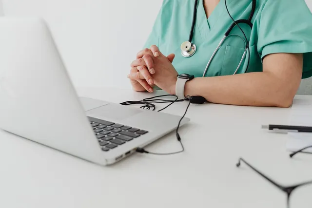 O persoană în uniformă medicală verde stă la o masă albă lângă un laptop deschis și un stetoscoap.