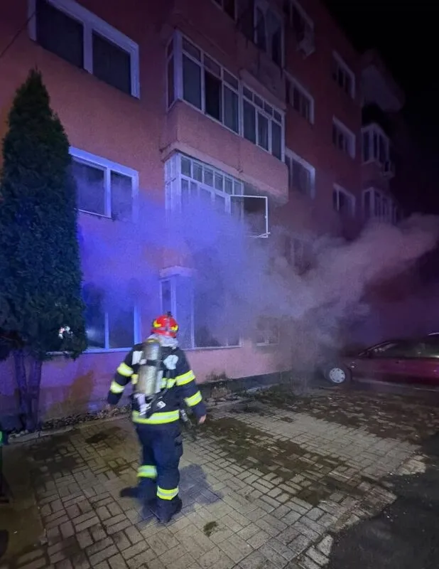 Un pompier cu echipament de protecție și butelie de oxigen stă pe un trotuar pavat lângă un bloc de locuințe din care ies fum.