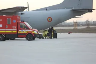 O mașină de ambulanță roșie și albă stă pe o pistă lângă o aeronavie gri mare, în prezența unor persoane și a unor bagaje.