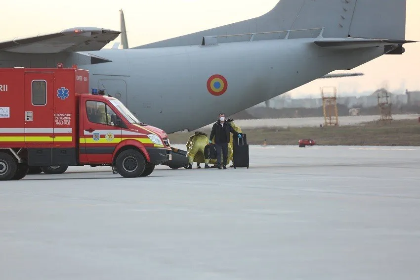O mașină de ambulanță roșie și albă stă pe o pistă lângă o aeronavie gri mare, în prezența unor persoane și a unor bagaje.