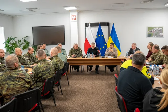 Un grup de persoane în uniformă militară și civilă stă la o masă lungă într-o încăpere cu steaguri și un ecran negru pe perete.