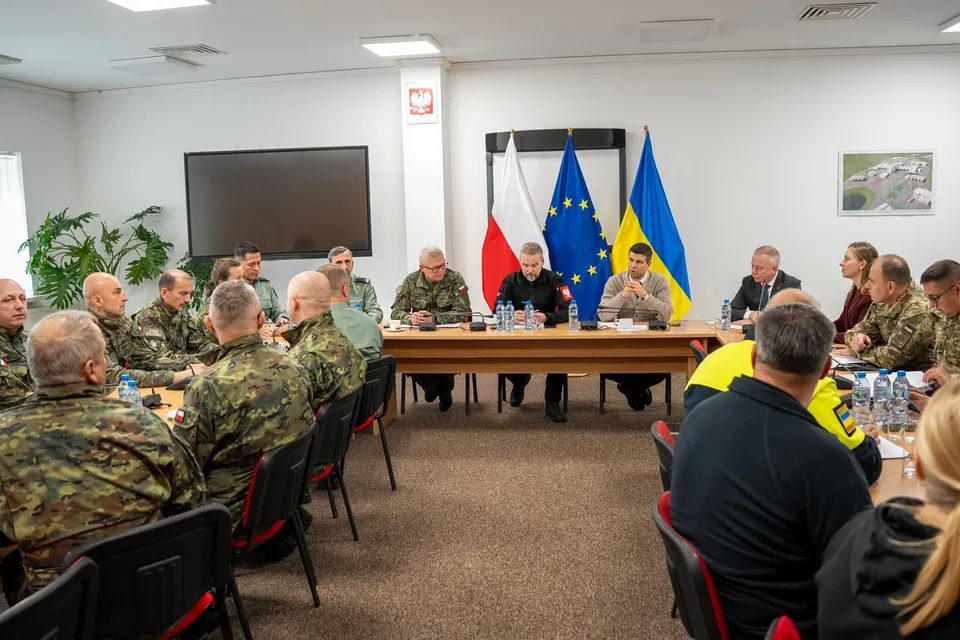 Un grup de persoane în uniformă militară și civilă stă la o masă lungă într-o încăpere cu steaguri și un ecran negru pe perete.