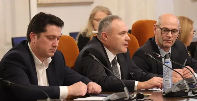 Trei bărbați în costume și cămași stau la o masă cu microfoane și o sticlă de apă în fața unui fundal de perete deschis.