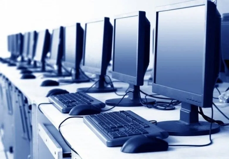 Un rând de monitoare de calculator, tastaturi și mouse-uri dispuse pe un birou alb într-o încăpere cu lumină albastră.