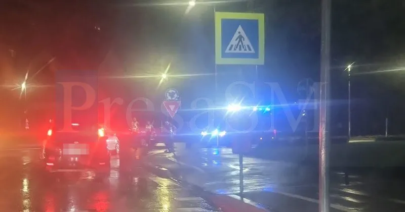 Autoturisme cu luminile aprinse stau pe un drum umed lângă un semn de trecere de pietoni sub lumină nocturnă.
