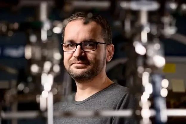 Un bărbat cu ochelari și barbă scurtă poartă un tricou gri în fața unor echipamente de laborator metalice și neclare.
