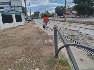 O persoană merge pe un drum de pământ și pietriș lângă șine de tramvai și un gard metalic gri.