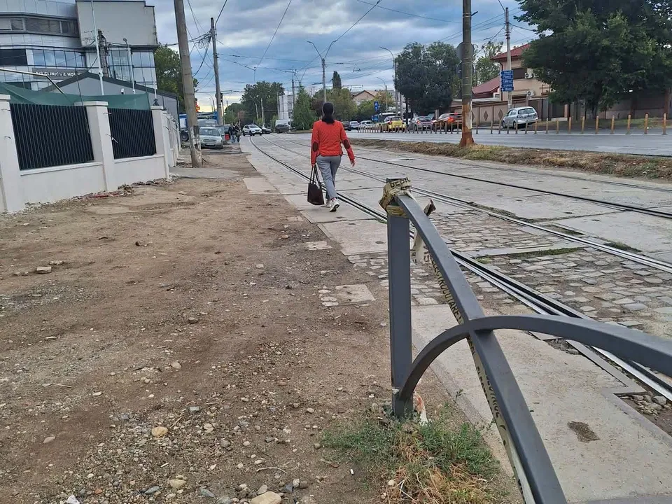 O persoană merge pe un drum de pământ și pietriș lângă șine de tramvai și un gard metalic gri.
