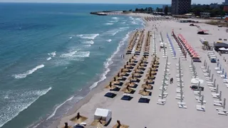 Aerial view of a sandy beach lined with rows of white and colored beach umbrellas next to the blue sea under a clear sky.