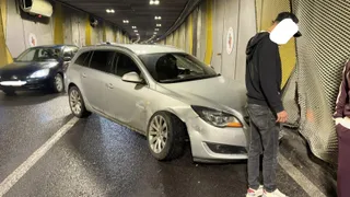 Un autoturism argintiu stă într-un tunel lângă un parapet, în timp ce un bărbat cu hanorac negru stă în picioare lângă o plasă metalică.