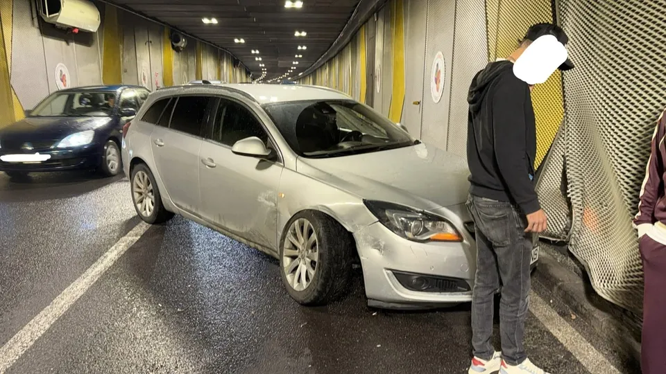 Un autoturism argintiu stă într-un tunel lângă un parapet, în timp ce un bărbat cu hanorac negru stă în picioare lângă o plasă metalică.