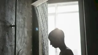 Silueta unei persoane stă sub un duș din care curge apă sub lumina unei ferestre mari.