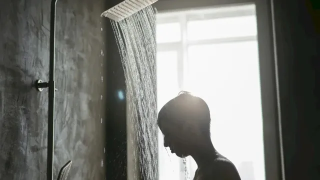 Silueta unei persoane stă sub un duș din care curge apă sub lumina unei ferestre mari.