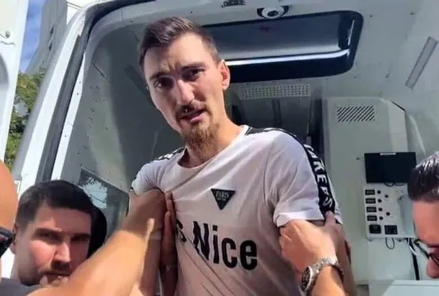 Un bărbat cu barbă scurtă poartă un tricou alb cu inscripții negre, fiind ținut de mai multe persoane într-un spațiu interior alb.