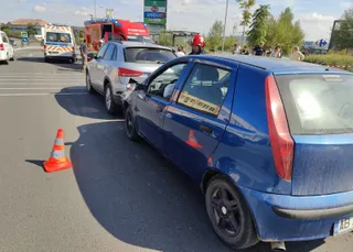 Un con traficului portocaliu stă pe asfalt lângă un autoturism alb și un autoturism albastru pe o stradă cu vehicule de serviciu în fundal.