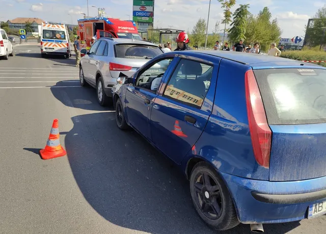 Un con traficului portocaliu stă pe asfalt lângă un autoturism alb și un autoturism albastru pe o stradă cu vehicule de serviciu în fundal.