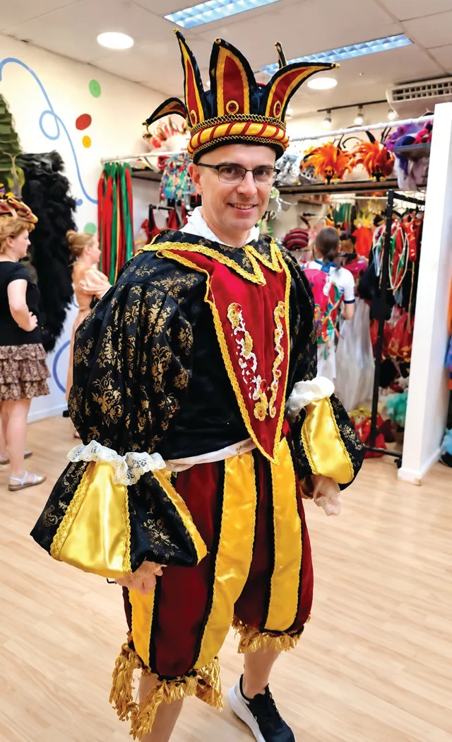 Un bărbat zâmbind poartă un costum de bufon cu pantaloni galbeni și roșii, într-un magazin cu costume de carnaval.