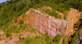 O stâncă mare de piatră cu suprafețe verticale este înconjurată de vegetație verde pe un versant de munte.