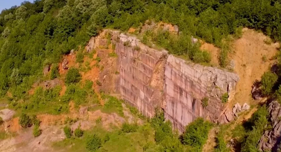 O stâncă mare de piatră cu suprafețe verticale este înconjurată de vegetație verde pe un versant de munte.