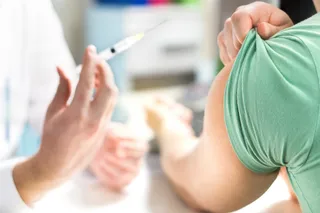 O persoană în halat alb ține o seringă cu lichid transparent lângă brațul unei persoane care poartă un tricou verde.