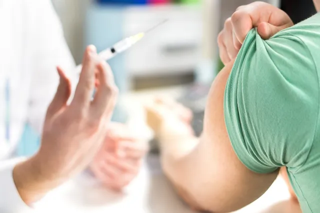 O persoană în halat alb ține o seringă cu lichid transparent lângă brațul unei persoane care poartă un tricou verde.