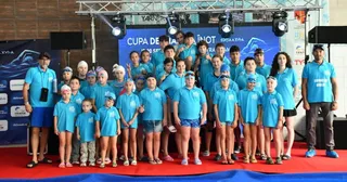 Un grup mare de copii și adulți în tricouri albastre stau în picioare pe un covor roșu în fața unui panou de fundal albastru.