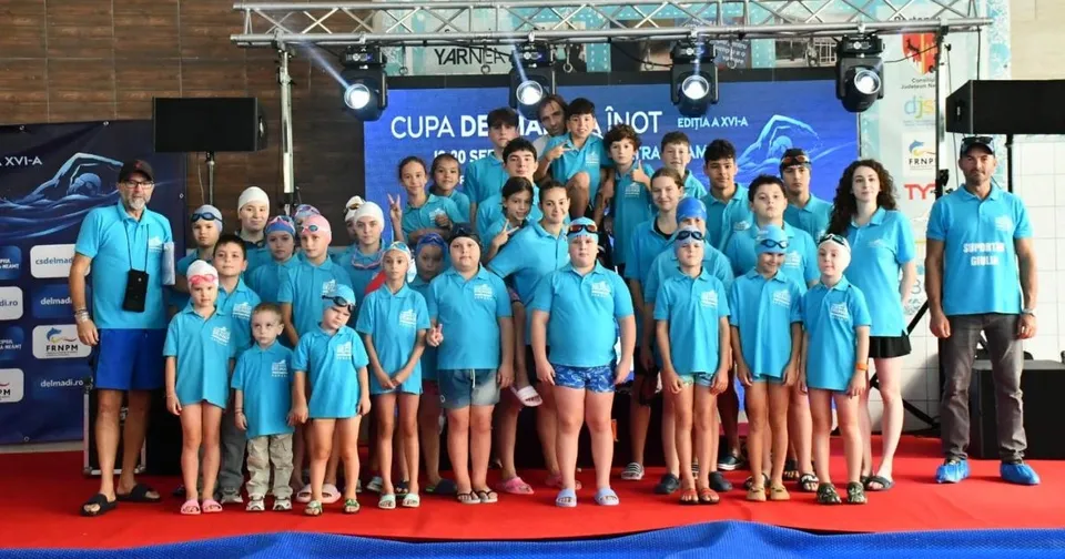 Un grup mare de copii și adulți în tricouri albastre stau în picioare pe un covor roșu în fața unui panou de fundal albastru.