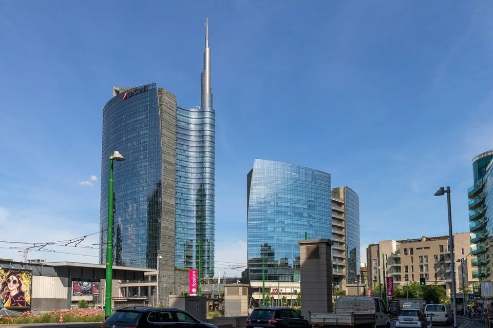 Mai multe clădiri înalte din sticlă se ridică sub un cer senin, lângă un drum cu mașini și stâlpi de iluminat public.