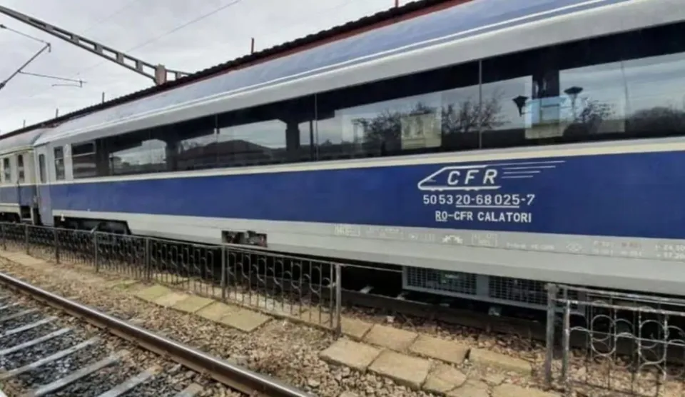 Un tren albastru și gri cu inscripția CFR rulează pe șine lângă un gard metalic negru.