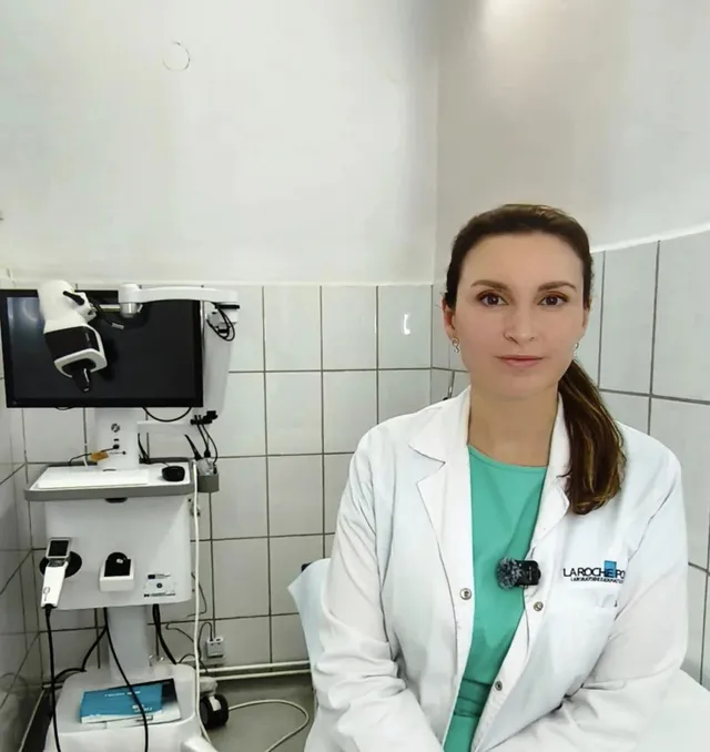 O femeie cu părul castaniu legat într-o coadă de cal poartă un halat alb și stă într-o cameră cu pereți albi plătiți.