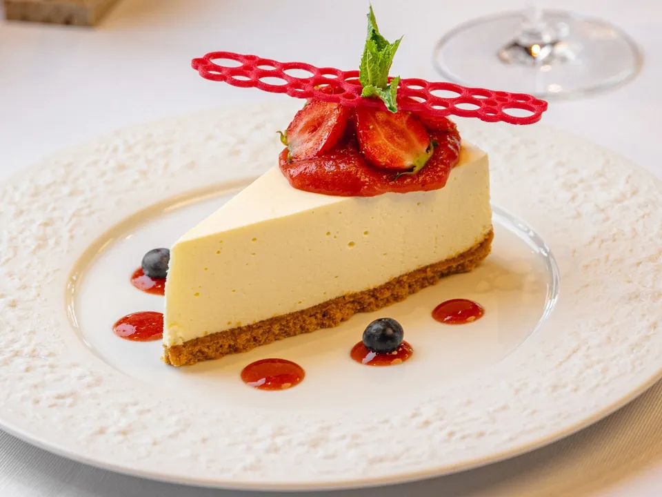 O felie de cheesecake cu fructe de pădure și decor de fructe roșii este servită pe o farfurie albă decorată.