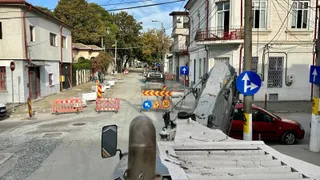 Un vehicul utilitar cu braț hidraulic este parcat pe un drum de oraș lângă un autoturism roșu și semne de circulație.