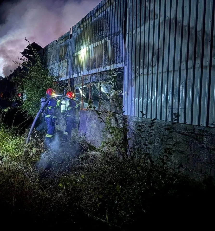 Două persoane purtând echipament de protecție și căști roșii folosesc un furtun pentru a stinge un incendiu la o clădire cu perete metalic striat.
