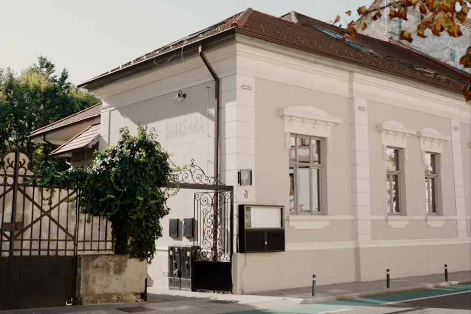 O clădire cu fațadă gri și albă, având ferestre înalte și un gard metalic negru lângă o tufă verde.