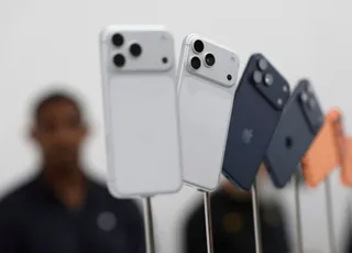 Patru telefoane inteligente sunt așezate vertical pe suporturi sub un fundal alb, cu o persoană neclară în planul secund.