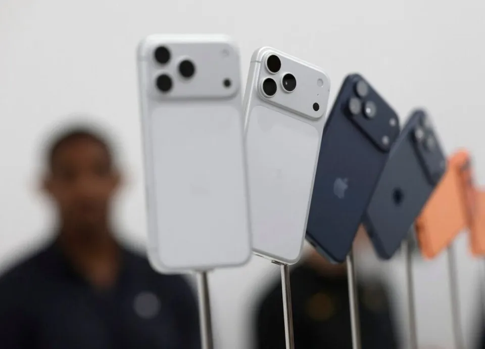 Patru telefoane inteligente sunt așezate vertical pe suporturi sub un fundal alb, cu o persoană neclară în planul secund.