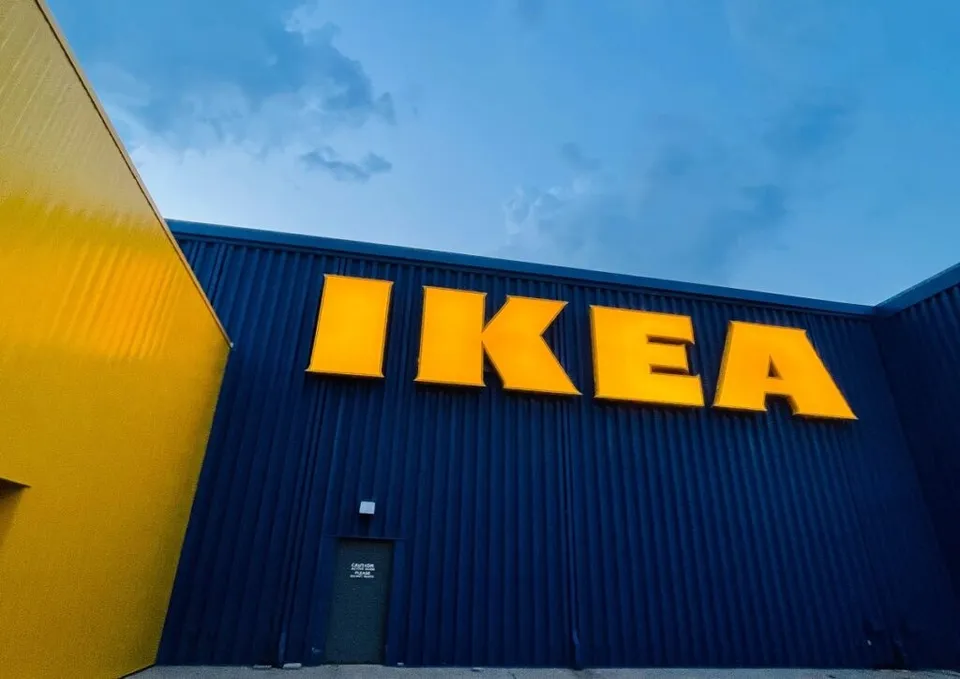 Un perete albastru cu textul galben IKEA este situat între o structură galbenă și un cer înnorat.