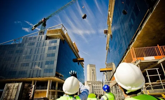 Oamenii de pe un șantier poartă căști de protecție albe și verzi sub o macara care ridică obiecte între două clădiri în construcție.