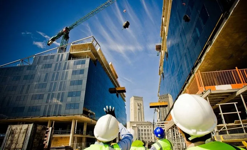 Oamenii de pe un șantier poartă căști de protecție albe și verzi sub o macara care ridică obiecte între două clădiri în construcție.