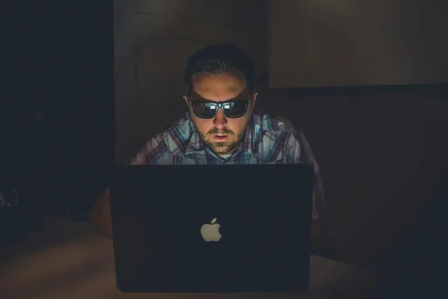 Un bărbat cu ochelari de soare stă la o masă și privește un laptop deschis într-un mediu întunecat.