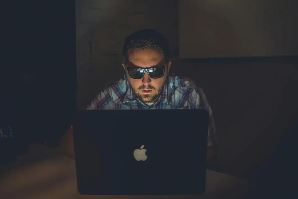 Un bărbat cu ochelari de soare stă la o masă și privește un laptop deschis într-un mediu întunecat.