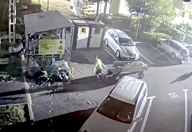 O persoană purtând o vestă reflectorizantă se află lângă un utilitar și un morman de resturi pe o suprafață asfaltată lângă câteva mașini parcate.
