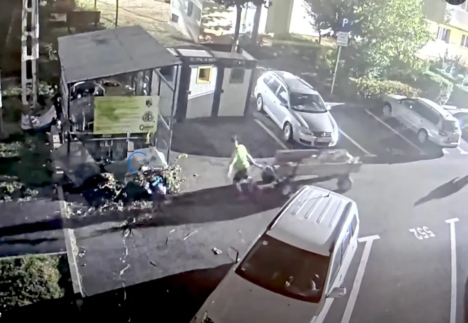 O persoană purtând o vestă reflectorizantă se află lângă un utilitar și un morman de resturi pe o suprafață asfaltată lângă câteva mașini parcate.